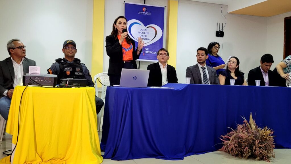 Mesa de autoridades de lançamento do projeto Quem escolhe o seu caminho? Você ou as Drogas? em Matões. De pé, a promotora de justiça Cristiane Lago aparece falando ao microfone atrás de uma mesa com toalha azul, na qual aparecem três homens e uma mulher. Do lado esquerdo, aparece outra mesa, com toalha amarela e dois homens sentados. Na parede ao fundo o banner do projeto e próximo a ele uma mulher de pé. 