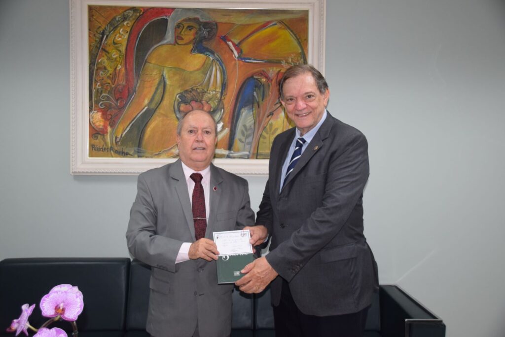 A imagem mostra o procurador-geral de justiça, Eduardo Nicolau recebendo um livro das mãos do advogado Sergio Tamer. Os dois estão de pé em frente a um sofá cinza e um quadro na parede. 