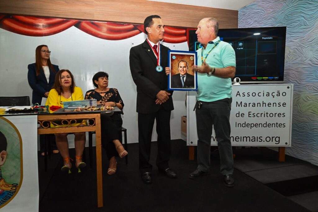 Procurador-geral de justiça, Eduardo Nicolau, recebe um quadro das mãos do presidente da Amclam, coronel Furtado. Na imagem, aparecem ainda três mulheres no lado esquerdo. Do lado direito, aparece um banner pequeno onde se lê Associação Maranhense de Escritores Independentes. 